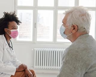 Young female doctor sitting with elderly patient. Both are wearing masks.