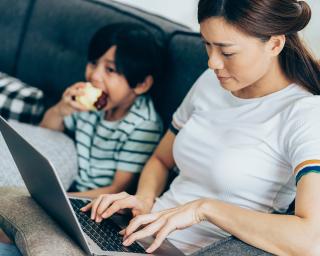 Young Asian American mother with young child sitting on couch next to her eating an apple, while she searches the internet on a laptop