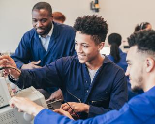 Two mixed race engineering students work on a group project together in a workshop whilst their tutor watches in the background. They are all wearing blue coveralls.