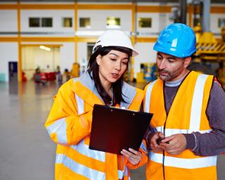 Shot of two warehouse workers talking together over a clipboard while standing inside of a large warehouse